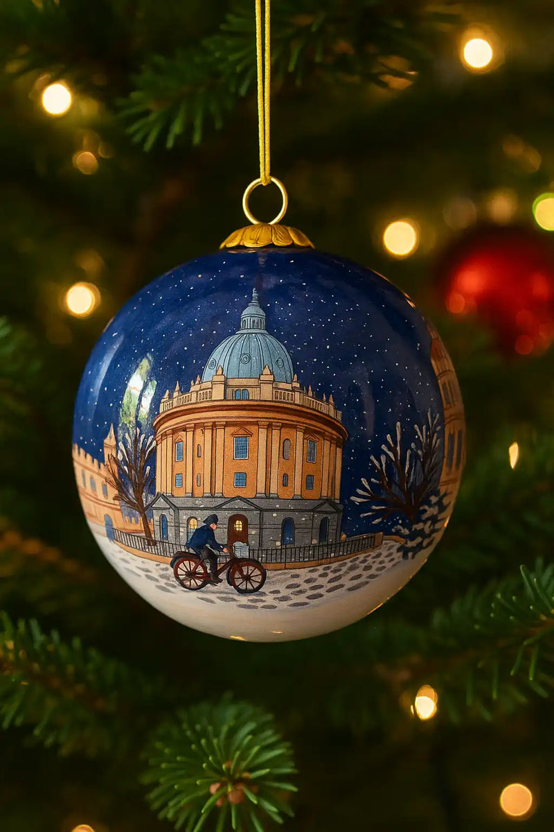 A blue Christmas ornament depicting a snowy Oxford landmark with a cyclist.