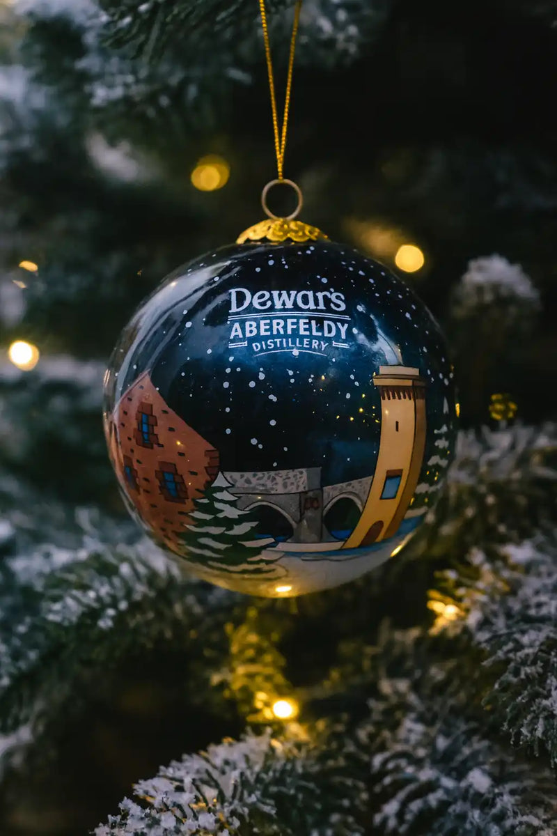 A dark blue, spherical Christmas ornament with a snowy landscape design and the text ’Dewar’s Aberfeldy Distillery’.