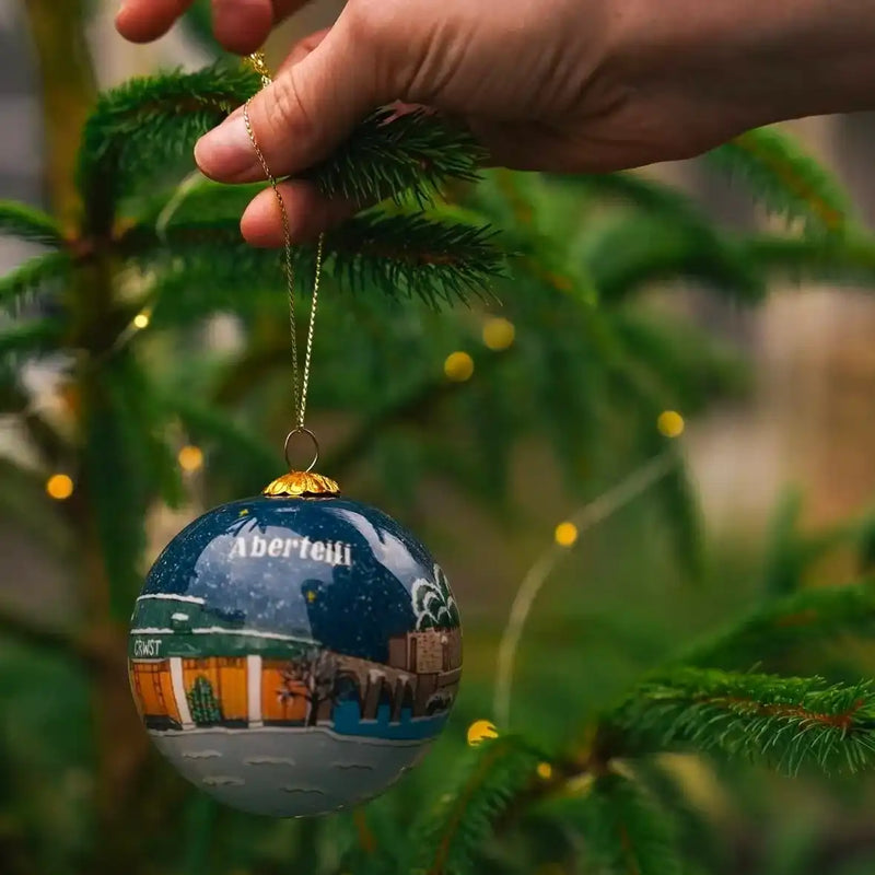 A hand-painted Christmas ornament depicting a winter landscape with buildings, hanging from a pine tree branch.