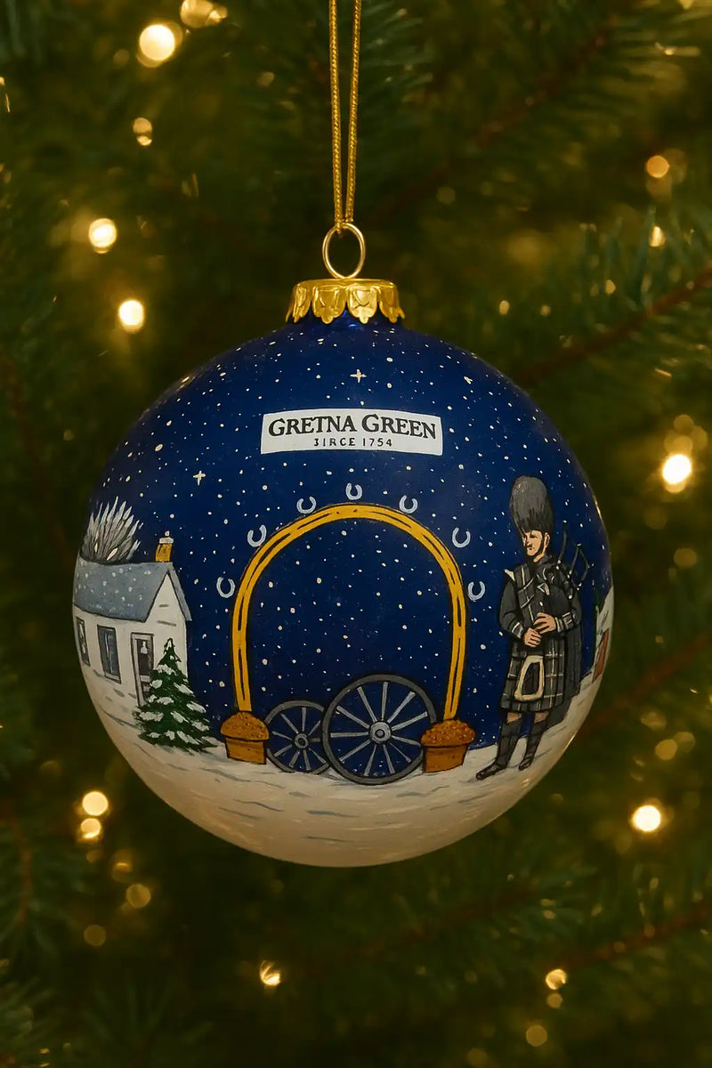 A navy blue Christmas ornament featuring a Scottish-themed winter scene with a building, a bagpiper, and an archway of horseshoes.